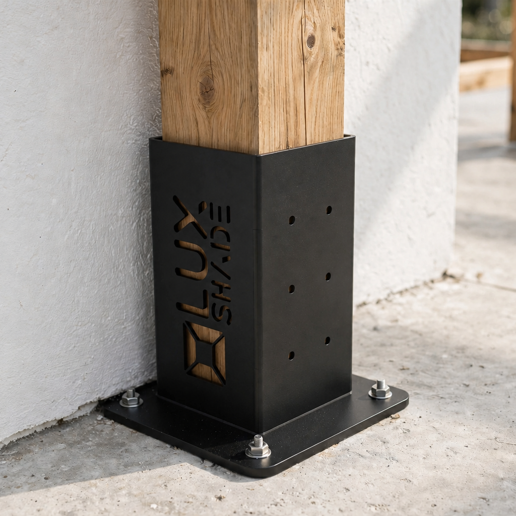A black metal post base bracket with the brand name 'LUX SHIELD' engraved vertically, shown installed outdoors against a white stucco wall with a wooden post passing through it.