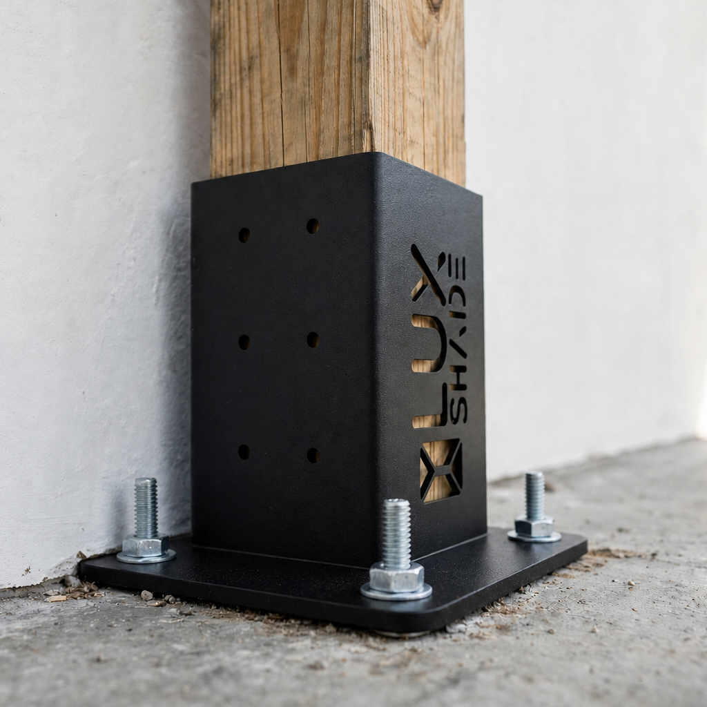 A black metal post base bracket with a wooden post inserted and four mounting bolts, photographed outdoors against a white wall and concrete ground. The side of the bracket has a cutout logo reading 'XJL'.