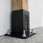 A black metal post base bracket with a wooden post inserted and four mounting bolts, photographed outdoors against a white wall and concrete ground. The side of the bracket has a cutout logo reading 'XJL'.