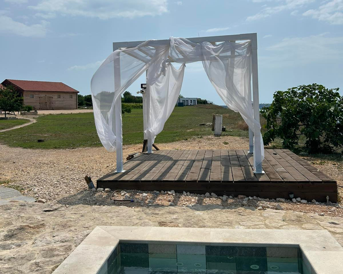 Wooden platform with white fabric draped over it, set against a scenic background with a building and greenery.