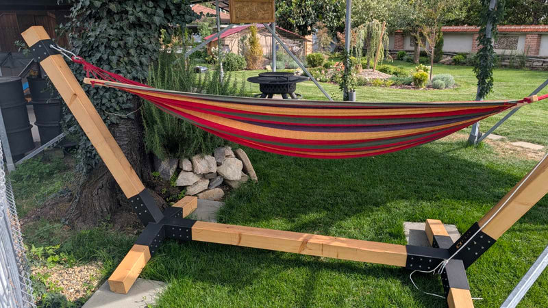 Hammock on a stand in a backyard setting with trees and rocks.